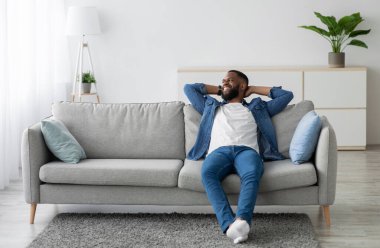 Neşeli, çekici, bin yıllık Afrikalı Amerikalı erkek koltukta oturmuş minimalist oturma odasındaki boşluğa bakıyor. Evde dinlen, mola ver. Erkekler hafta sonlarını ve boş zamanlarını severler.