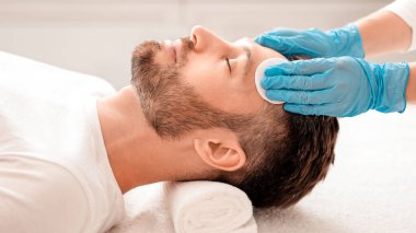 Orta yaşlı yakışıklı adam kozmetolojiste gidiyor, erkek yüz bakımı konsepti. Tanınmayan spa görevlisinin, tedavi öncesi pamuk pedi kullanarak sakallı adamı tonerle temizleyişinin yan görüntüsü.