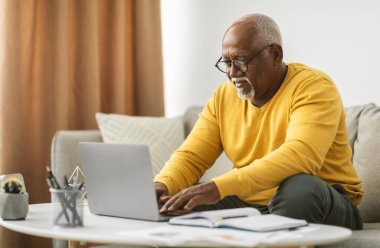 Üst düzey Black Freelancer, dizüstü bilgisayarda çalışıyor, bilgisayarla çalışıyor, içeride koltukta oturuyor. Evde internete göz gezdiren, gözlük takan bir erkek. Serbest Kariyer Konsepti