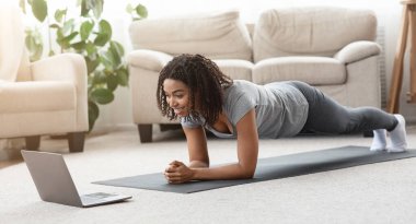 Evde antrenman. Siyah bir kadın dizüstü bilgisayar başında yoga yaparken, egzersiz yaparken, oturma odasında, boş alanda, panoramada...