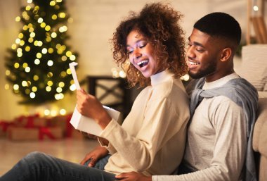 Heyecanlı Afro-Amerikalı çift Noel ağacının önünde oturma odasında Noel hediyesini açıyor.