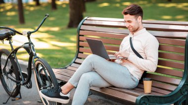 Mola sırasında dışarıda çalış. Çekici adam bankta oturur, bilgisayarda kahve ve bisikletle yazar, boşluğu kopyalardı.