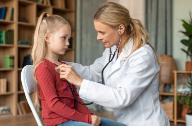 Kadın doktor, evdeki sağlık kontrolü sırasında çocuğun ciğerlerini kontrol ediyor. Dost canlısı bayan çocuk doktoru steteskop kullanarak genç bir hastanın nefes alışını ve kalp atışlarını inceliyor.
