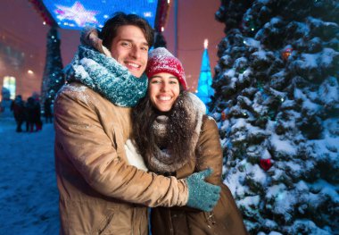 Şehirde Kış Randevusu. Neşeli çift kameraya gülümsüyor Noel ağacının yanında duruyor geceleri dışarıda yürüyor