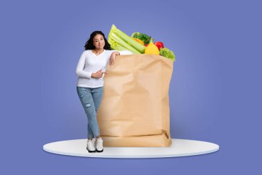 Bir müşteri taze meyve ve sebzelerle dolu büyük bir kesekağıdının yanında duruyor. Arka plan basit bir mavi, yeni normal ürünler almanın zevkini vurguluyor..