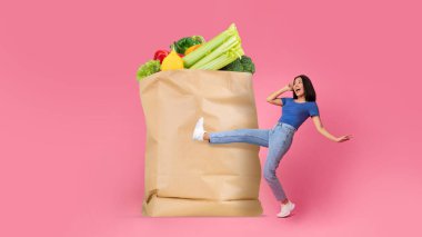 Neşeli bir müşteri renkli yiyeceklerle dolu büyük bir kese kağıdının yanında duruyor. Arka plan canlı pembe, modern bir yaşam tarzında taze yiyecek alışverişinin neşesini vurguluyor..
