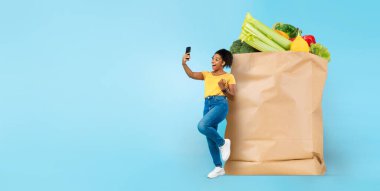 Neşeli bir müşteri, içi taze yiyeceklerle dolu büyük bir kesekağıdının yanında duruyor. Akıllı bir telefonu var ve gülümsüyor. Modern alışverişin rahatlığını kucaklıyor..