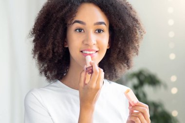 Kadın sabah makyajı için hazırlanıyor, balsam yapıyor, evdeki banyoya koyuyor. Neşeli genç Afro-Amerikalı bayan dudaklarına nemlendirici ruj sürüyor aynaya bakıyor, boş yer var.