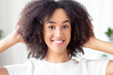 Kadın güzelliği, modern blog yazarı ve çevrimiçi yaşam korsanları. Neşeli, Afro-Amerikan bayan ellerini kıvırcık, sağlıklı saçlara bastırıyor ve oturma odasının iç tarafındaki kameraya bakıyor.