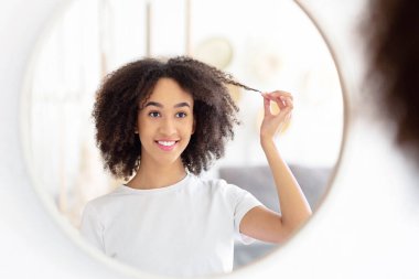 Tatlı bir kadının yuvarlak aynasında yansıması, güzellik bakımı, tavsiye ve modern blog yazarının reklamı. Genç gülümseyen Afrikalı Amerikalı kadın sabah oturma odasında kıvırcık saçlı bir kilit tutuyor.