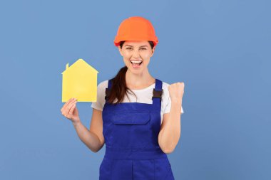 Aşırı neşeli Sert Şapkalı Genç Kadın ve Kapaklı Evinde Sarı Kağıtlar Taşıyor, Başarılı Yenilemeyi Kutluyor Mavi Stüdyo Arkaplanında Duran Neşeli Yakışıklı Kadın, Boşluğu Kopyala