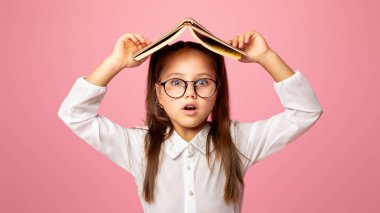 Derslerden bıktım. Gözlüklü ilkokul kızı kafasını kitapla örtüyor, pembe arka planda soyutlanmış.