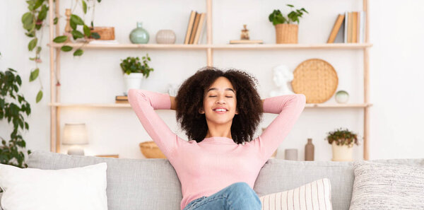 Наконец, один дома, чистота в квартире и человеческие эмоции. Счастливая молодая африканская американка с закрытыми глазами наслаждается тишиной, бросает руки за голову, сидит на диване в интерьере гостиной