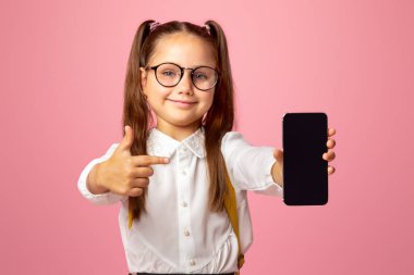 İncelenecek malzemeler. Gözlüklü liseli kız, okul üniformalı, pembe arka planda izole edilmiş, boş bir akıllı telefon ekranında parmağını gösteriyor.