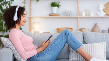 İş, evde eğitim ve kovid 19 tecridi sırasında online müzik. Gülümseyen Afro-Amerikan kadın kulaklıklı, kulaklıklı, koltukta oturan ve oturma odasında telefonla yazan.