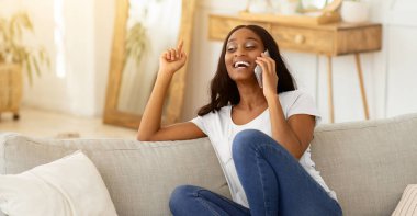 Çekici Afrikalı Amerikalı kadın akıllı telefondan konuşuyor ve evdeki kanepede gülüyor. Güzel siyahi bayan, arkadaşlarıyla kapalı mekanda harika bir konuşma yapıyor.