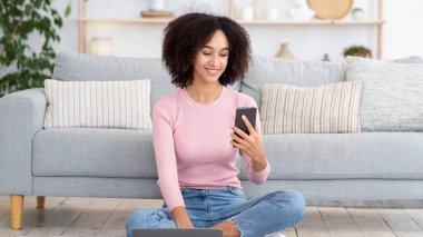 Menajer evden çalışır, müşteri araması yapar ve sosyal uzaklık sırasında modern bir iş yapar. Neşeli genç Afro-Amerikan kadın dizüstü bilgisayarı ve fincanı ile yerde oturuyor ve rahat oturma odasında telefonda yazıyor.