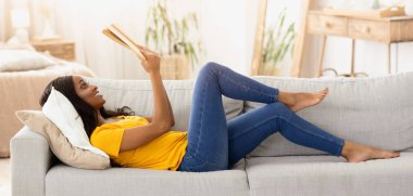 Evde hobilerin olsun. Milenyum siyahi kadın kanepeye uzanmış, kitap açık, yan görüş. Genç Afrikalı Amerikalı kadın oturma odasında heyecan verici bir hikaye okuyor.