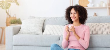 Ev hapsi süresince evde çalışmadan önce rahatlayın ve kahve için boş zamanınız olsun. Neşeli bin yıllık Afro-Amerikan kadın sıcak bir bardak sıcak içecekle oturma odasında, yerde oturuyor.