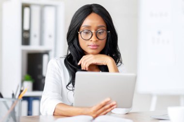 Modern ofisindeki iş yerinde tablet tableti kullanan odaklanmış siyahi bir iş kadını. Çevrimiçi önemli belgeyi okuyan, e-postaları kontrol eden, iş konferansı veren kendinden emin bir girişimci.