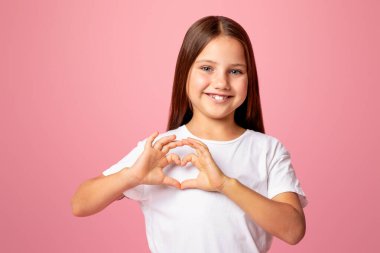 Sevgi ve şefkat duygularının ifadesi. Gülümseyen küçük kız elleriyle kalp işareti gösteriyor, pembe arka planda izole edilmiş, stüdyo çekimlerinde.
