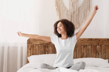 Günaydın, yeni gün, hafta sonu, tatil. Mutlu Afro-Amerikan zindelik timsali kadın yatakta oturur, uyuduktan sonra kollarını uzatır ve rahat yatak odasının içinde, rahat bir sabah geçirir.