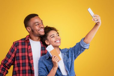 Afrika kökenli Amerikalı eşler telefonda, seyahat biletleri ve sarı arka planda duran bavullarla selfie çekiyorlar. Tatil, Tatil ve Turizm Konsepti İçin Hazır