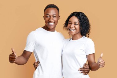 Sarılıp sarılan iki mutlu zenci sevgili sarı stüdyo arka planında baş parmağını gösteriyor. Neşeli Afro-Amerikalı kadın ve erkek kucaklaşıp el kol hareketi yapıyorlar, ürün, hizmet veya alım öneriyorlar.