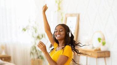 Kulaklığı ve cep telefonu olan mutlu siyah kadın evde en sevdiği müzikle dans ediyor. Afro-Amerikalı kadın güzel bir şarkıya taşınıyor, harika vakit geçiriyor.