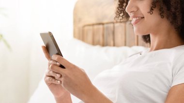 Video görüşmesi, online sohbet, sosyal ağlar ve COVID-19 salgını sırasında evde internette sörf. Gülümseyen Afro-Amerikan kadın yatakta uzanıyor ve telefonda yazıyor, yatak odasında, iç mekanda, yan bakışta.
