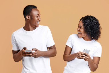 Joyful young african american man and woman playing together and competing in video games on smartphones, yellow studio background. Emotional black couple playing online games together, having fun