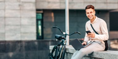 Modern dijital aletlerle dışarıda çalış. Dizüstü bilgisayarı, akıllı telefonu ve kulaklığı olan gülümseyen genç işadamı bisiklet yakınında oturuyor.