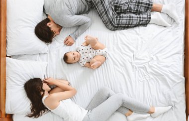 Genç bir aile yatakta. Küçük bebek yatağın ortasında yatıyor, ebeveynler yan yana uyuyor.