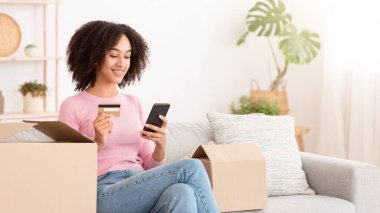 Ev teslimatı, sipariş ödeme, para ve modern alışveriş. Gülümseyen Afro-Amerikan kadın akıllı telefonuyla yazı yazıyor, kredi kartı tutuyor, oturma odasındaki kanepede oturuyor, boş yer var.