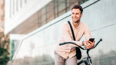Şehir caddesinde bisikletinin yanında duran genç bir adam gülümseyerek telefonunu kontrol ediyor. Hafif kıyafetler giyip, günün tadını çıkarırken rahat bir şehir yaşam tarzını somutlaştırıyor.