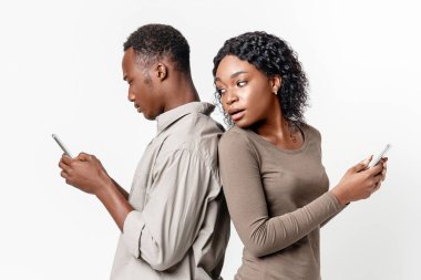 Sırt sırta verip gri stüdyo arka planında akıllı telefon kullanan siyah bir çift. Afrikalı Amerikalı kadın erkek arkadaşını gözetliyor sosyal medyada oturuyor, cep telefonu tutuyor, boş yer var.