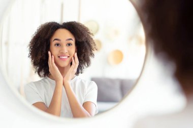 Evdeki spa gününden sonra sağlıklı cilt ve sağlık hizmetleri. Milenyum Gülümseyen Afrikalı Amerikalı kadın ellerini yanaklarına bastırıyor ve oturma odasının iç tarafındaki yuvarlak aynada yansımasından zevk alıyor.