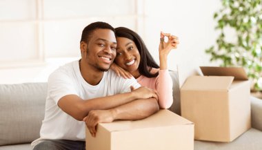 Ev anahtarı ve karton kutuları olan mutlu siyah çift kameraya bakıyor ve mülklerinde gülümsüyor, panorama. Genç Afrikalı Amerikalı ev sahipleri yeni bir daireye taşınacakları için heyecanlı.