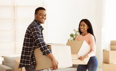 Siyah milenyum çifti taşınma gününde yeni dairelerinde karton kutular taşıyor. Eşyaları olan Afrikalı Amerikalı ev sahipleri gülümseyip kameraya, manzaraya bakıyorlar.