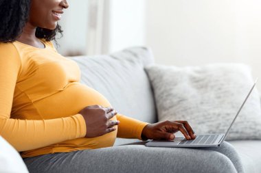 Evde dizüstü bilgisayar kullanan, oturma odasındaki koltukta oturan, boş bir alanda gülümseyen zenci hamile kadının yan görüntüsü. Mutlu Afro-Amerikan kadınları internette sörf bekliyor, annelik hakkında blog okuyor.