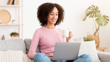 Covid-19 tecridi sırasında yeni normal, çevrimiçi uzaktan kumanda, eğitim, Webinar. Neşeli, Afro-Amerikalı bir kadın bardaktan su içiyor, kanepeye oturuyor, dizüstü bilgisayarla oturma odasında oturuyor ve düşünüyor.