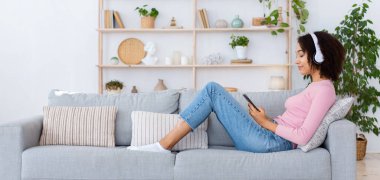 Boş zaman, hafta sonları, COVID-19 salgını sırasında evde cihazlarla dinlenmek. Sıradan giyinmiş gülümseyen Afro-Amerikan bayan, kulaklıklı koltukta, akıllı telefonuyla oturma odasındaki müziğin keyfini çıkarıyor.