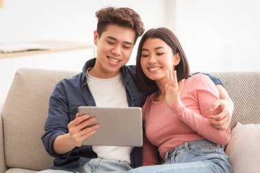 Dijital tabletli Asyalı bir çift evdeki koltukta oturan web kamerasına el sallayarak görüntülü arama yapıyor. Çin aile videosu bilgisayarı arıyor. Modern Uzaktan İletişim Konsepti