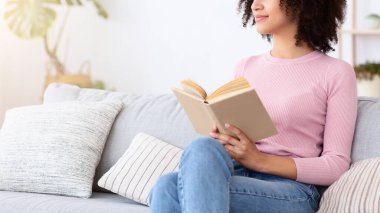 Serene oldukça genç Afro-Amerikan kadını kendini rahat hissediyor, bayan rüyası rahat olmanın keyfini çıkarıyor modern oturma odasındaki rahat havayı soluyor, kitap okuyor ve pencereden dışarı bakıyor, bedava uzay.