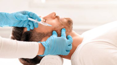 Sakallı, orta yaşlı bir adam güzellik salonunda güzellik iğnesi yaptırıyor. Kozmetik uzmanı klinikteki yakışıklı adam için yaşlanma önleyici prosedür uygulaması yapan mavi lastik eldivenleri teslim etti. Yüz bakımı, erkek kozmetoloji konsepti.