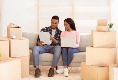 Genç siyah çift dizüstü bilgisayarda yeni evleri için tasarım projesi yapıyor. Ev dekorasyonu ve mobilya fikirleri için interneti karıştıran belgeleri olan genç bayan ve kocası şaşırdılar.