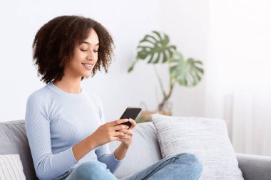 Sohbet, blogda sörf, modern sosyal ağlar, mobil uygulamalar. Bin yıllık Afro-Amerikan kadın akıllı telefonuyla kanepede oturuyor, oturma odasında mesaj yazıyor, boş yer var.