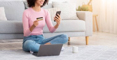 Çevrimiçi alışveriş, sipariş malları, karantinada çalışırken kredi kartıyla ödeme. Neşeli, Afro-Amerikalı genç bayan elinde bir fincan kakao ve dizüstü bilgisayarla oturmuş oturma odasında telefonla yazı yazıyor.