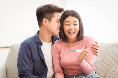 Pozitif Hamilelik Testi. Neşeli Asyalı koca ve eş, kutlama sonuçlarına sarılıyor, evde koltukta oturan bebeği bekliyor. Mutlu Ebeveynler ve Çocuk Doğumu Konsepti. Seçici Odaklanma