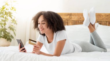 Cihazlı alımlar, COVID-19 salgını sırasında evde ödeme yapmak için mobil uygulama. Neşeli, güzel, zoomer, Afro-Amerikan kadın. Yatağa uzanmış, akıllı telefona bakıyor, kredi kartı tutuyor, yatak odasının içinde.